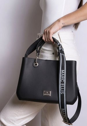 Vrouw houdt zwarte Marc Ellis-handtas met afneembare merkriem vast, draagt witte broek en wit mouwloos topje, zijaanzicht.