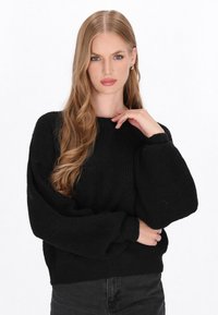 Pulover negru din tricot cu o textură striată, având un guler rotund și mâneci supradimensionate. Purtați cu pantaloni negri, modelul își ține o mână la tâmplă, în mod gânditor.