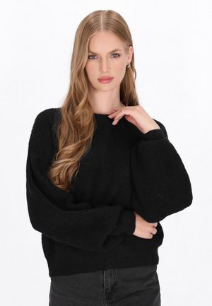 Schwarzer Strickpullover mit gerippter Textur, rundem Ausschnitt und übergroßen Ärmeln. Kombiniert mit schwarzen Hosen hält das Model eine Hand nachdenklich.