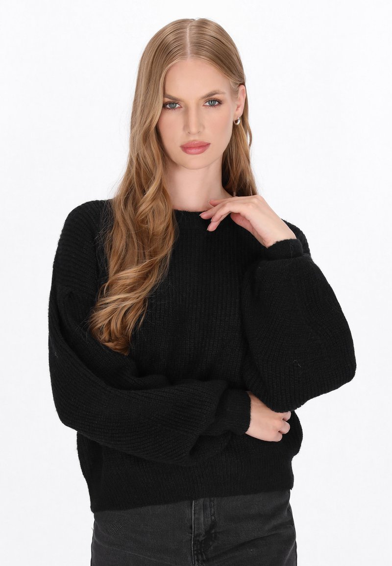 Zwarte gebreide trui met een geribbelde textuur, met een ronde halslijn en oversized mouwen. Draag deze met zwarte broek, terwijl het model met één hand nadenkend poseert.
