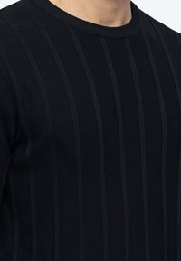 Μαύρο πουλόβερ με κάθετες ριγωτές ρίγες, που φοριέται από ένα άτομο, δείχνοντας την περιοχή πάνω από το στήθος και τον λαιμό, με φωτεινό φόντο.