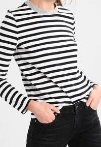 Zwart-wit gestreept shirt met lange mouwen en een grijze halslijn. Zachte, aansluitende stof. Draagt een high-waisted zwarte jeans.