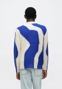 Suéter azul y crema con un patrón ondulado, que presenta un acabado texturizado y puños acanalados. La vista trasera muestra un contraste de color prominente y costuras visibles.