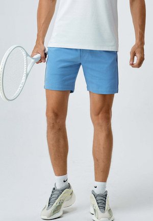 Mann trägt blaue Sportshorts, weiße Socken und graue Turnschuhe und hält einen weißen Tennisschläger in einer Hand.