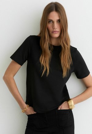 Kobieta o długich jasnobrązowych włosach, ubrana w czarną koszulkę z krótkim rękawem i czarne spodnie, z rękami w kieszeniach, nosząca złotą bransoletkę i masywną opaskę.