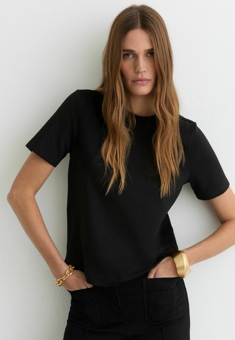 Nainen, jolla on pitkät vaaleanruskeat hiukset, musta lyhythihainen paita ja mustat housut, kädet taskuissa, kultainen rannekoru ja paksu kalvosin.