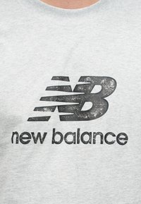 Φθαρμένο γκρι μπλουζάκι με φθαρμένο μαύρο λογότυπο New Balance που απεικονίζει τα στιλιζαρισμένα γράμματα "NB" και το όνομα της μάρκας από κάτω.
