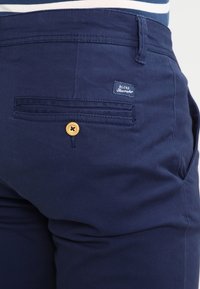 Námořnické modré kalhoty se zadní kapsou s zlatým knoflíkem a textilním štítkem, na kterém je napsáno "BLEND Seammaker." Hladká, strukturovaná látka.