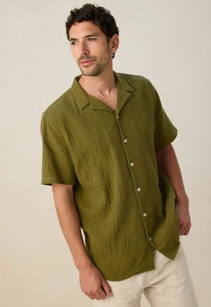 Chemise olive à manches courtes avec un motif texturé, fermeture à bouton et col à revers, associée à un pantalon de couleur claire.