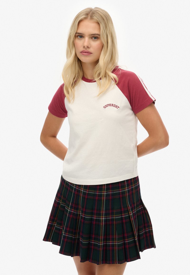 Camiseta blanca con mangas raglán en color granate y el logo "Superdry". Combinada con una falda de cuadros plisada en verde oscuro, rojo, amarillo y blanco.