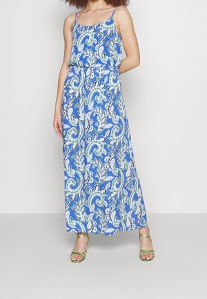 Nainen pukeutunut siniseen, hihattomaan maximekkoon, jossa valkoinen ja vihreä paisley-kuvio, yhdistettynä metallinvihreisiin remmikenkiin.