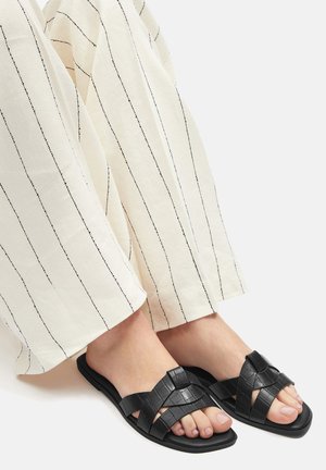 Sandalias sintéticas negras con un diseño trenzado, con una suela plana. Combinadas con pantalones de pierna ancha a rayas blancas.