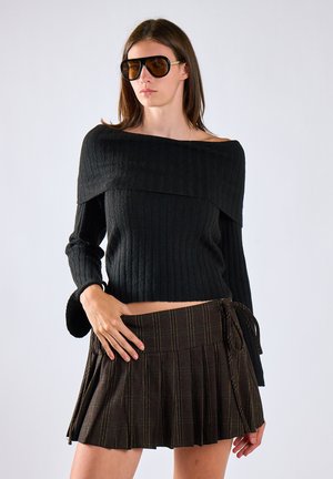 Mujer con gafas de sol marrones de gran tamaño, suéter negro acanalado sin hombros y falda corta plisada a cuadros marrón oscuro con lazo lateral.