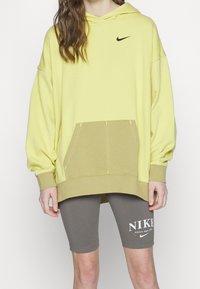 Hoodie giallo oversize con logo Nike nero, dotato di tasca frontale e accenti beige-verdastri a contrasto. Abbinato a pantaloni corti grigi.
