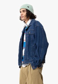 Blå denimjacka med framfickor, urblekt finish och knappstängning. Buren över en flerfärgad randig skjorta och matchad med khakibyxor.