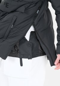 Svart isolerad jacka med dragkedja och knappstängning, elastiska ärmslut och justerbar nederkant. Fodrad insida synlig med samlad tyg.