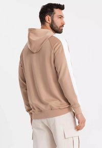Felpa beige con cappuccio e strisce laterali bianche, realizzata in tessuto morbido, con una vestibilità comoda e polsini a costine. Indossata con pantaloni chiari.