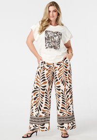 Chemise blanche à manches courtes avec un imprimé botanique, associée à un pantalon large couleur crème, aux motifs de feuilles brunes et oranges, et des sandales noires.