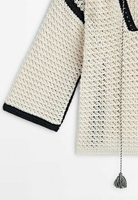 Haut au crochet en blanc cassé avec des accents noirs. Présente une coupe ample, un col en V et un pendentif décoratif. Design texturé en tricot ouvert.
