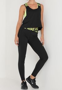 Top corto sportivo nero con un accento giallo neon, abbinato a leggings neri. Presenta il marchio "NIKE PRO". Indossato con scarpe sportive nere.