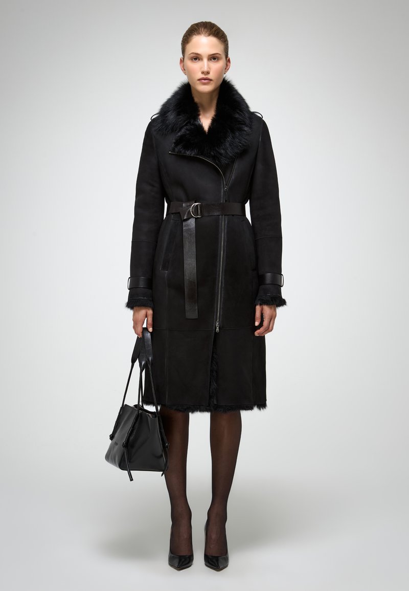 Mustat mokkasatiinista valmistettu takki, jossa on keinoturkiskaulus, vyöllinen vyötärö ja vetoketjullinen etuosa. Puvussa on turkkisaumoja hihansuissa ja helman kohdalla. Malli pitää mustaa laukkua.