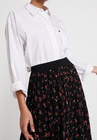 Vit skjorta med rullade ärmar, kombinerad med en svart plisserad kjol med ett blommönster i röd och grön samt ett elastiskt midjeband.