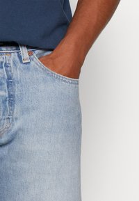 Jeans de mezclilla azul claro con corte recto, que presentan un bolsillo frontal y un sutil desgastado en la tela. Accesorios de metal visibles.