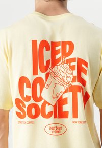 Camiseta amarilla claro con texto rojo en negrita: "SOCIEDAD DEL CAFÉ HELADO." Incluye un gráfico de una taza de café con crema batida.