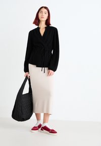Cardigan nero avvolgente con cintura e maniche ampie, abbinato a una gonna midi beige. Una borsa tote nera trapuntata e sneakers rosse con dettagli bianchi completano il look.