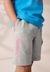 Pantaloni scurți gri din bumbac, cu imprimeu roz strălucitor "SUBSCRIBE" pe piciorul stâng. Asortați cu o cămașă albastră cu mânecă scurtă, cu croială lejeră.