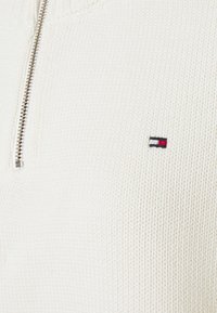 Weiße gestrickte Zip-Up-Strickjacke mit hohem Kragen. Hat ein kleines, mehrfarbiges Logo auf der Brust und einen strukturierten Stoff. Silberner Reißverschluss.