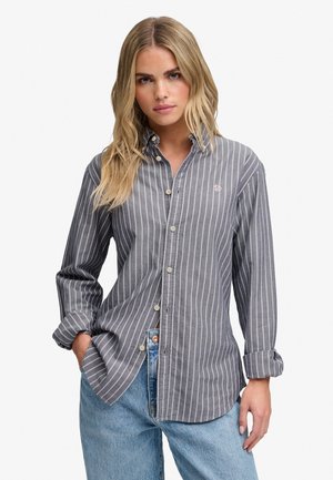 Kvinde med langt blondt hår iført en grå langærmet skjorte med hvide lodrette striber og lyseblå jeans.