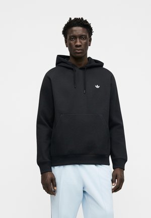 Mężczyzna ubrany w czarną bluzę z kapturem z małym białym logo Adidas na piersi i jasnoniebieskie spodnie, stojący na jednolitym tle.