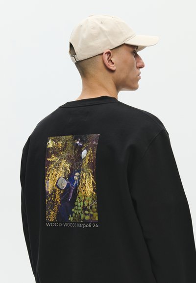 Hombre con gorra beige y sudadera negra con diseño en la parte trasera de flores amarillas y hojas verdes, texto "WOOD WOOD Marpoli 26".