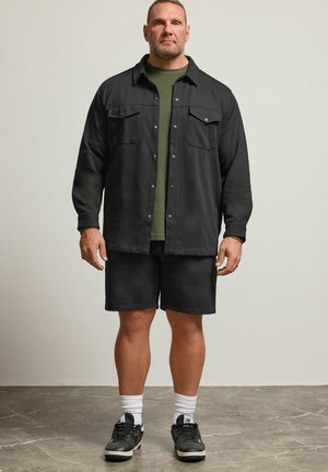 Mies seisoo musta nappijakun, oliivinvihreän paidan, mustien shortsejen, valkoisten sukkien ja mustien lenkkareiden kanssa harmaalla lattialla.