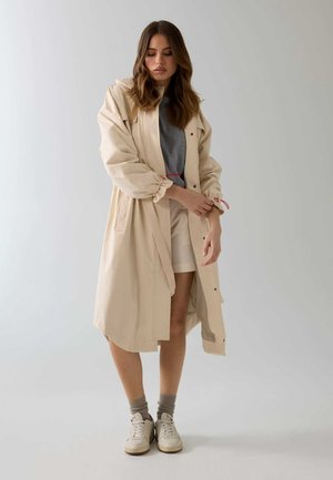 Femme ajustant la manche d'un long trench-coat beige, portant une chemise grise, un short blanc, des chaussettes grises et des baskets blanches, debout devant un fond uni.