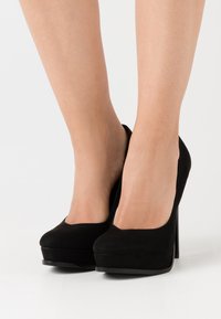 Svart mocka högklackade skor med plattformdesign, rund tå och en slank, glänsande stilettklack, som visar en mjuk textur.