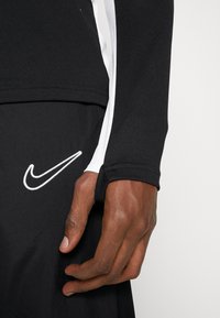 Black athletic top with long sleeves, featuring white accents and a Nike logo. Smooth fabric texture and fitted design. Hand visible on the left.
