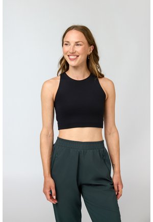 Smilende kvinne med langt hår iført svart ermeløs crop top og mørkegrønne bukser med elastisk midje mot ensfarget bakgrunn.