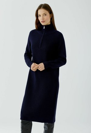 Frau, die ein langes marineblaues Strickkleid mit Halb-Reißverschluss-Kragen und schwarzen kniehohen Stiefeln trägt, steht vor einem einfarbigen Hintergrund.