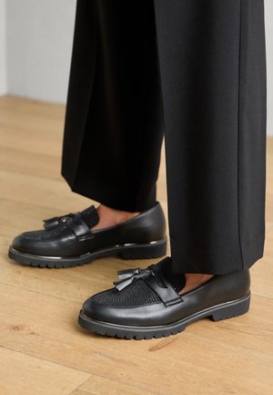 Chaussures en cuir noir avec un design perforé, semelle plate et détail de noeud décoratif, assorties à un pantalon noir ample.