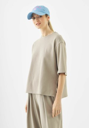 Jonge vrouw met een losse beige outfit en een lichtblauwe baseballpet, staand tegen een effen witte achtergrond met een lichte glimlach.