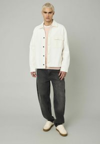Jeune homme aux cheveux blonds courts portant une veste blanche, une chemise rose clair, un jean noir ample et des baskets blanches, se tenant devant un fond uni.