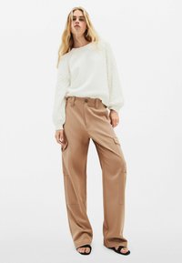 Maglione bianco con maniche a trecce texture, abbinato a pantaloni cargo marroni a gamba larga con tasche laterali e chiusura con bottone.