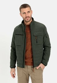 Quiltet mørkegrøn jakke med høj krave, flere lommer og lynlås. Båret over en tekstureret orange sweater.