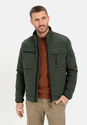 Quilted dark green jacket with a high collar, multiple pockets, and a zip closure. Worn over a textured orange sweater.