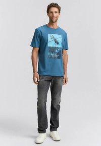 Žalsvai mėlyna trumpomis rankovėmis marškinėliai su grafiniu atvaizdu, vaizduojančiu sraigtasparnį, derinami su tamsiais džinsais ir baltais sportbačiais. Medvilnės medžiaga, laisvas stilius.