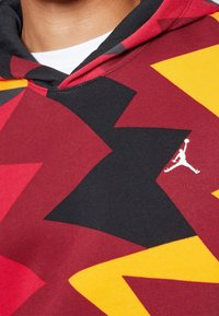 Sweat à capuche avec un motif géométrique audacieux en rouge, noir et jaune, arborant un logo Jumpman blanc sur le devant. Texture de tissu doux.
