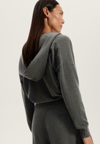 Hanorac gri cu glugă, cu o croială lejeră și manșete cu nervuri, având o textură moale și un design minimal. Viziunea din spate arată detaliul glugii.