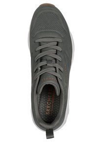 Zapatilla deportiva gris con una parte superior perforada, cordones texturizados y una suela blanca. Presenta un logo y una tira en el talón.